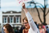 A woman holds up a sign that reads “Which of My Students Do I Shield With My Body?” Photo credit Heather Mount/Unsplash