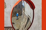 Backpacker with a bright blue backpack’s reflection in a round mirror, is seen walking down a medieval street, away from the camera. There is an orange sun visor on the mirror and that is reflected in an orange frame around the photo.