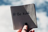 A hand holding a napkin with the hashtag “Be Kind” written on it