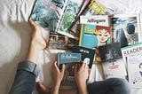 View of person sitting on bed reading, all you see are the lower halves of their legs, clad in blue jeans and with bare feet. On the bed there are several magazines and books spread around for the reader to look at.