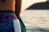 A woman sitting on a beach, staring out over a blue ocean with a bottle of sunscreen lying against her leg.