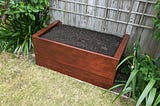 Brown raised box filled with compost and soil next to a fence and a lawn