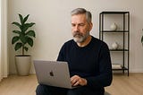 A designer in his 60s sitting on a pouf while typing on a MacBook