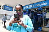 A woman operating a TECNO smartphone outside a store. TECNO, itel, and Infinix are owned by Transsion