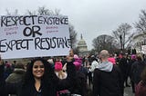 Photo of Emmeline Rodriguez holding a sign saying ‘Respect Existence or Expect Resistance’ in English with smaller red lettering reading ‘Respecto Salud / Dignidad Justicia’ in spanish. She is on the street joined by other protesters near the Capitol building in Washington D.C. All people in the photo wear warm clothing, including Emmeline who wears a black jacket and black scarf.