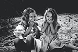 black and white image of two young girls maybe 5–6 years old, sitting on a piece of driftwood on a sandy beach. Both girls are laughing/smiling and covering their mouths with both palms. Girl on left is wearing white striped dress, girl on right is wearing grey dress. shortsleeves.