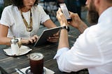 Two people sit across from each other at a wooden cafe table, one working on a laptop and the other on a cell phone.