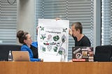 Two people in a business room setting. A woman in a blue jacket, a man with a black shirt, holding a paper with suggestions of change mobility for the better.
