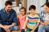 A man, a woman, a girl, and a boy all sat on sofa and looking at an Amazon Echo device