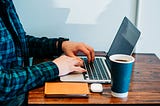 This image shows a person sitting at a wooden table, typing on a laptop.