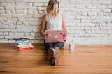 Lady sitting on a wooden floor next to a pile of books carrying a pink laptop.