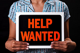 Woman holding a black sign which reads help wanted in red letters.