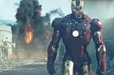 Iron Man’s gold-and-red suit, on the right side of the image, appears to be walking away from an explosion (orange flames and black smoke) in a sandy, barren area.
