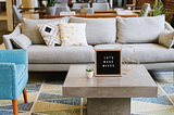 A coworking space featuring a couch, an armchair, and a concrete coffee table with a sign reading, “let’s make waves” atop it.