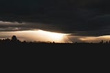 Photo of horizon of golden light between  black silhouette bands of landscape with distant mountains and heavy cloud above.