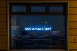 Photo of a lit neon sign “What is Your Story?” in a dark window.