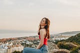 woman sitting on a rock overlooking a beautiful village, she’s in a red top and jeans, long auburn hair