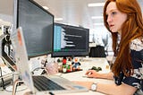 A woman with long red hair is sitting at a desk working on two computer screens and a laptop.