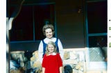A woman and a young girl are standing together in front of a house with a stone facade. The girl is wearing a bright red dress with a white collar and the woman is dressed in a white blouse with a blue jumper.