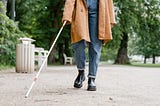 A blind person walking in a park using their white cane. Only the lower half of the person is visible, with the white cane in focus.