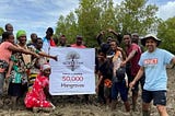 A group of people standing happily around a banner that reads “join us in planting 50,000 mangroves.”