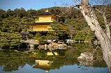 Kinkaku-ji (Golden Pavilion) in Kyoto, Japan, a three-story Zen Buddhist temple with gold-leaf-covered upper floors, reflected in a tranquil pond and surrounded by lush greenery. A large tree trunk appears in the foreground, adding depth to the serene landscape.