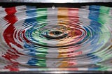 A droplet breaks a colorful surface of water and causes a ripple effect outwards.
