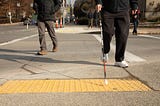 Two people crossing an intersection with the image taken from a low angle showing the curb ramp. One person uses an assistive can typically used by people with visual impairments.