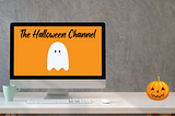 White desk in front of gray background. Computer monitor on desk shows The Halloween Channel title and logo ghost on orange background. Jack ‘O Lantern sits on desk.