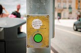 A green street crosswalk button for pedestrians, with a sticker placed above it that reads “Push to reset the world”.