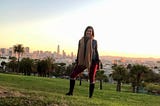 The author, a young woman, stands in a tree-lined park smiling. The San Francisco, California skyline and a sunset fill the background. She’s wearing a tan scarf, jean jacket, and pink yoga pants.