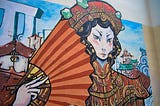 image features a colorful mural painting on a wall, depicting a stylized female figure dressed in traditional Chinese attire. The woman has an intense gaze, pale skin, and dark, wavy hair adorned with an ornate red and gold headdress decorated with green gemstones. She holds a large, red fan with sunburst-like stripes in her right hand. The mural also includes architectural elements resembling pagodas and classical Chinese buildings in the background, along with a blue sky and abstract structure