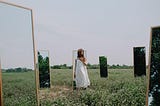 a photo of a Southeast Asian woman standing in a grass field surrounded by six haphazardly placed dressing mirrors