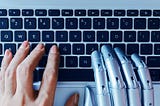 A human hand and a cyborg hand typing on a keyboard.