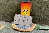 A phone with a sad face emoji is propped against a wall. A cardboard sign in front of it reads “Alms for a charger?” The phone rests on a knitted mat with a small candle next to it.