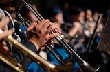 A picture of an orchestral brass section. The photo zooms in on a trumpet player’s hands. In the blurred background, you can see the french horn section.