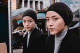 Woman with black head covering and jacket, and grey jersey top covering her neck, standing before a mirror which also shows her reflection. The reflection looks directly at the viewer which is incongruent with the woman outside her reflection, suggesting editing tricks/artwork to give this effect. Background is a street possibly in East Asia or China with an open air stall in the mirror reflection.