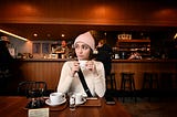 white woman with short brown hair, pink knitted cap, white shirt, holding small porcelain coffee cup in a café, looking away from the camera