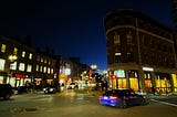 Street scene at night. Non-descript buildings with their lights on and cars passing on the street, a few pedestrians are walking on sidewalks.