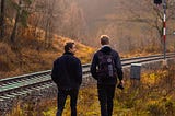 Two men in a rural environment are seen from the back, walking at the edges of a railway track while the traffic light is red