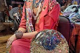 A shop window mannequin, her lovely female form adorned in coral beaded and bejeweled finery against a background of magnificent costumery. In the foreground sits a glittering crystal ball filled with colors