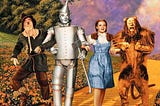 Movie still from The Wizard of Oz with the scarecrow, tin man, Dorothy, and the lion on the yellow brick road