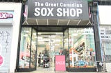 Storefront on a medium wide shot, with the store name ‘The Great Canaidan Sox Shop’. The store is open with the lights on and colourful displays seen through the window. A sign reading ‘SALE’ is placed just in front of the door.