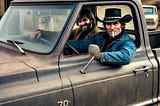 Two rowdy cowboys in a 1970s pickup truck with hats and cigarette