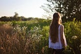 A young woman with long hair stands in alone in a meadow, her back to camera