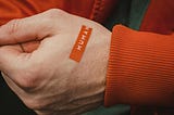 Closeup of a person’s hand with white skin that has an orange sticker on it which reads “human.” The person is wearing an orange coat with a gree shirt underneath.