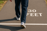 A person walks along a clearly marked 30-foot path on a sunlit paved trail, starting at the “0” line and heading toward the “30 FEET” mark. Only their lower body is visible, emphasizing motion and measurement.