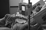 In the foreground, an IV machine, unfocused. In the background, a patient lying in a hospital bed, turned towards a window.