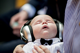 Close up photo of a baby in a white dress shirt and black bow tie with large headphones on asleep in the arms of an adult.