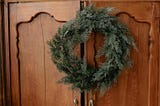 An image showing a rustic wooden cabinet with carved details on its doors. Hanging on the cabinet is a natural-looking evergreen wreath made of lush, green foliage, giving a festive or seasonal feel. The wood of the cabinet has a warm brown tone, and two metallic tassel-like ornaments with gold accents hang from the bottom of the wreath.